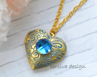 May include: A gold-colored heart-shaped locket necklace with blue filigree and a large, faceted blue gemstone. The necklace has a delicate gold chain. The text "corsiva design" is visible on the bottom right of the locket.