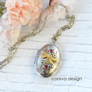 Puede incluir: Collar con dije ovalado de plata con un diseño de libélula dorada y detalles de piedras preciosas rojas. El dije tiene un borde adornado con marcas de números romanos. El collar está en una cadena de plata.