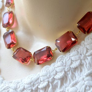 Puede incluir: Un collar con piedras rectangulares de cristal rojo oscuro facetado, engarzadas en metal de tono dorado. Las piedras están unidas en una fila continua, creando un diseño geométrico llamativo que descansa sobre una tela blanca texturizada.