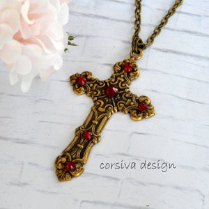 Puede incluir: Un collar con un colgante de cruz en tono dorado con acentos de pedrería roja. La cruz es adornada y detallada, con un diseño de filigrana. El collar está en una cadena.