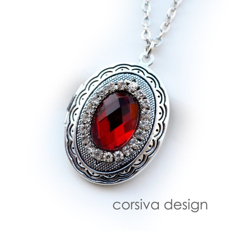May include: Silver locket necklace with a red gemstone surrounded by clear rhinestones. The locket has a detailed ornate design.