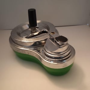Pode incluir: Um cinzeiro vintage com um design cromado e verde. O cinzeiro tem um topo circular com uma pega preta e uma base verde. O cinzeiro é feito de metal e tem uma estética retro.