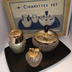 Pode incluir: Conjunto de cigarros vintage com cinzeiro, isqueiro e recipiente com detalhes em metal dourado. A caixa diz "Cigarettes Set". As peças estão em uma bandeja preta.