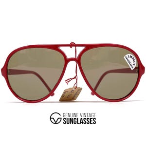 May include: Red vintage sunglasses with a classic aviator shape. The sunglasses have brown lenses and a small tag attached. The frame is a bright red color. The sunglasses have a sticker that says "Impact Resistant".