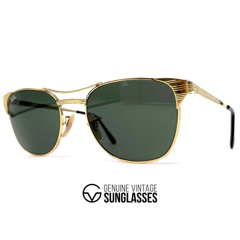 Op de afbeelding: Goudkleurige metalen zonnebril met donkergroene glazen en een dubbele neusbrug. Het montuur heeft gestructureerde details bij de veren en zwarte uiteinden. De tekst GENUINE VINTAGE SUNGLASSES staat onder de zonnebril afgedrukt.