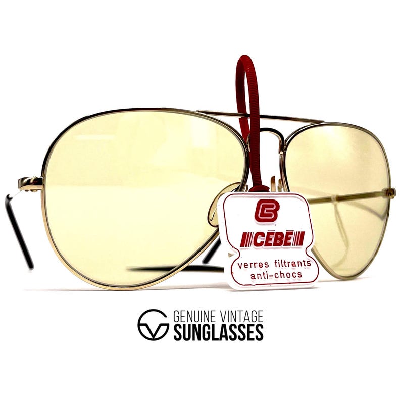 May include: Vintage aviator sunglasses with gold frames and yellow-tinted lenses. The glasses have a double bridge and black temples. A red tag with the brand name "CEBE" is attached. The text "GENUINE VINTAGE SUNGLASSES" is printed below.
