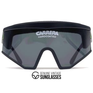 Puede incluir: Gafas de sol Carrera negras con lentes grises. Las gafas de sol tienen el nombre de la marca "Carrera Hardcoated" en blanco. La parte inferior de las gafas de sol tiene un diseño angular único. El texto "Genuine Vintage Sunglasses" está en la parte inferior.