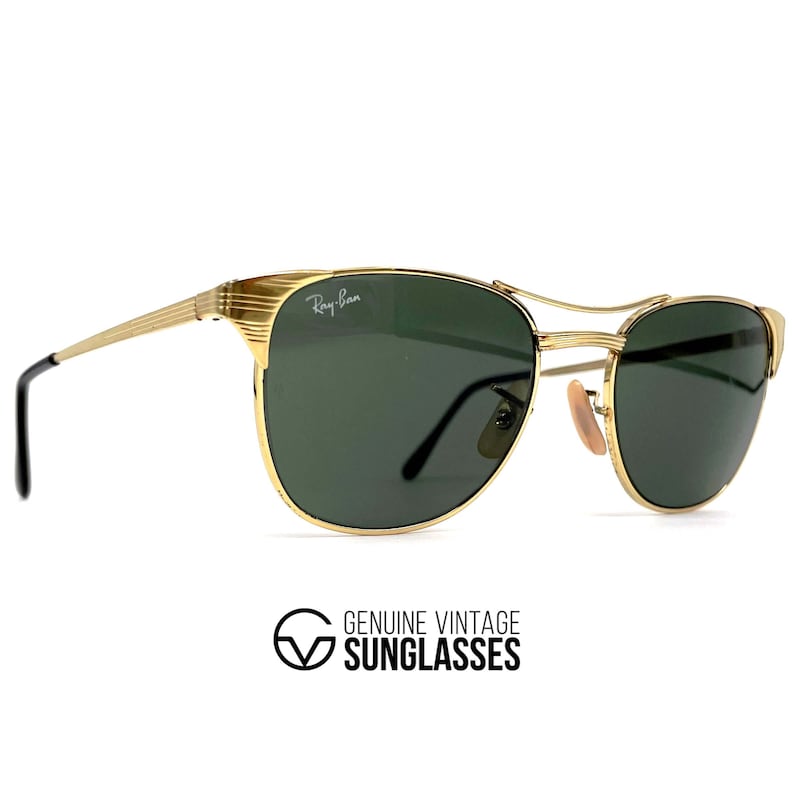 Op de afbeelding: Goudkleurige metalen zonnebril met donkergroene glazen, voorzien van het Ray-Ban logo op het rechterbovenglas. Het montuur heeft een dubbele brug, gestructureerde details op de veren en zwarte kunststof uiteinden voor extra comfort.