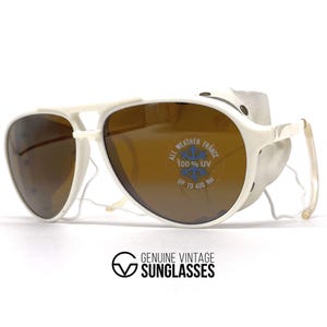 May include: White vintage sunglasses with brown lenses and side shields. The sunglasses have a white frame and the text "ALL WEATHER FRANCE 100% UV UP TO 400 NM" is visible on the lens. The bottom of the image has the text "GENUINE VINTAGE SUNGLASSES".