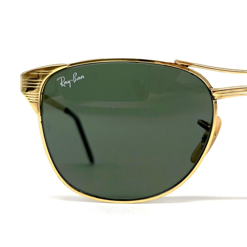 Op de afbeelding: Goudkleurige metalen zonnebril met donkergroene glazen en het witte Ray-Ban logo in de linkerbovenhoek. Het montuur is voorzien van een opvallend geribbeld detail bij het scharnier en een dunne constructie met een dubbele neusbrug.