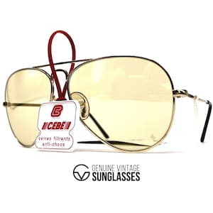 May include: Vintage aviator sunglasses with yellow-tinted lenses and a gold-colored metal frame. The sunglasses have a double bridge and black temples. A tag with the brand name "CEBE" is attached. The text "GENUINE VINTAGE SUNGLASSES" is also visible.
