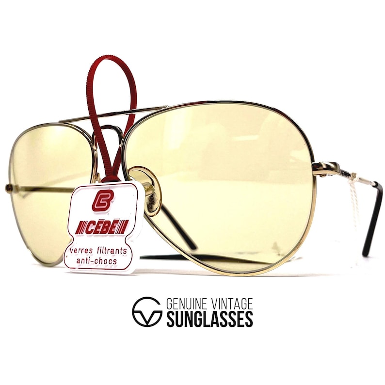 May include: Vintage aviator sunglasses with yellow-tinted lenses and a gold-colored metal frame. The sunglasses have a double bridge and black temples. A tag with the brand name "CEBE" is attached. The text "GENUINE VINTAGE SUNGLASSES" is also visible.
