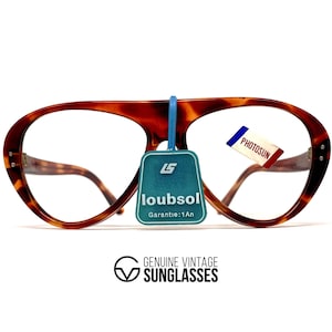 May include: Vintage tortoiseshell eyeglasses with a distinctive shape. The frames have a warm brown and amber pattern. A blue tag reads "loubsol Garantie:1An" and a small tag with "PHOTOSUN" is attached. The text "GENUINE VINTAGE SUNGLASSES" is below.