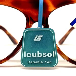 May include: Close-up of eyeglasses with a tortoiseshell pattern frame. A teal tag with the text "loubsol" and "Garantie: 1 An" is attached to the glasses. The tag also features a white logo.