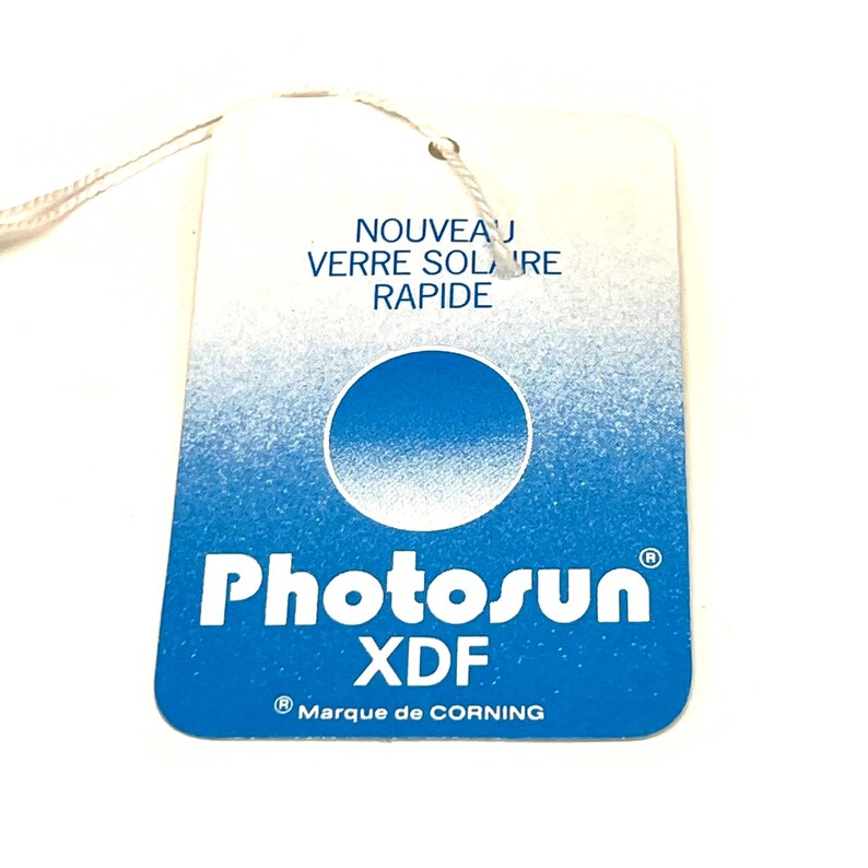 May include: A rectangular tag with a blue and white gradient background. The tag features the text "NOUVEAU VERRE SOLAIRE RAPIDE" and the brand name "Photosun XDF" in white. A blue circle is in the center of the tag.