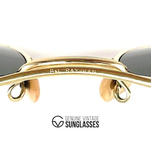 Op de afbeelding: Goudkleurige metalen zonnebril met een dubbele brug en de gegraveerde tekst B&L RAY-BAN. Het montuur bevat twee donkere glazen en twee beige neuspads. Onder het montuur staat de tekst GENUINE VINTAGE SUNGLASSES met een bijbehorend logo.