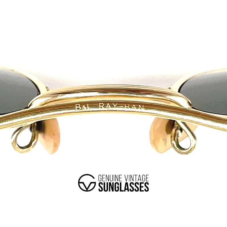 Op de afbeelding: Goudkleurige metalen zonnebril met een dubbele brug en de gegraveerde tekst B&L RAY-BAN. Het montuur bevat twee donkere glazen en twee beige neuspads. Onder het montuur staat de tekst GENUINE VINTAGE SUNGLASSES met een bijbehorend logo.