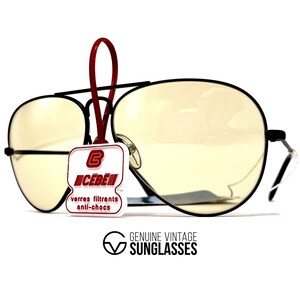 Puede incluir: Gafas de sol de aviador con montura negra y lentes de color amarillo. Una etiqueta roja cuelga de la montura, mostrando el logotipo y el texto de la marca. La parte inferior de la imagen contiene el texto "GENUINE VINTAGE SUNGLASSES".