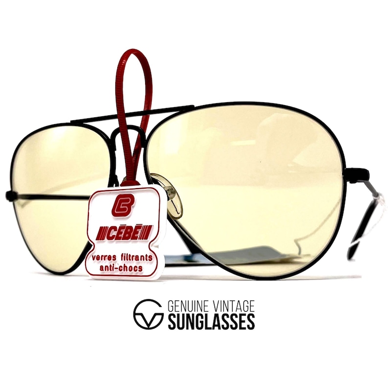 Puede incluir: Gafas de sol de aviador con montura negra y lentes de color amarillo. Una etiqueta roja cuelga de la montura, mostrando el logotipo y el texto de la marca. La parte inferior de la imagen contiene el texto "GENUINE VINTAGE SUNGLASSES".