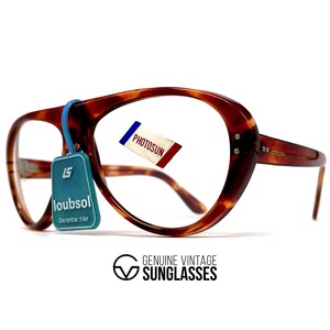 May include: Vintage tortoiseshell eyeglasses with a large, rounded frame. The glasses have a blue tag that reads "loubsol" and a small tag that reads "PHOTOSUN". The frame is brown with a marbled pattern. The text "GENUINE VINTAGE SUNGLASSES" is at the bottom.