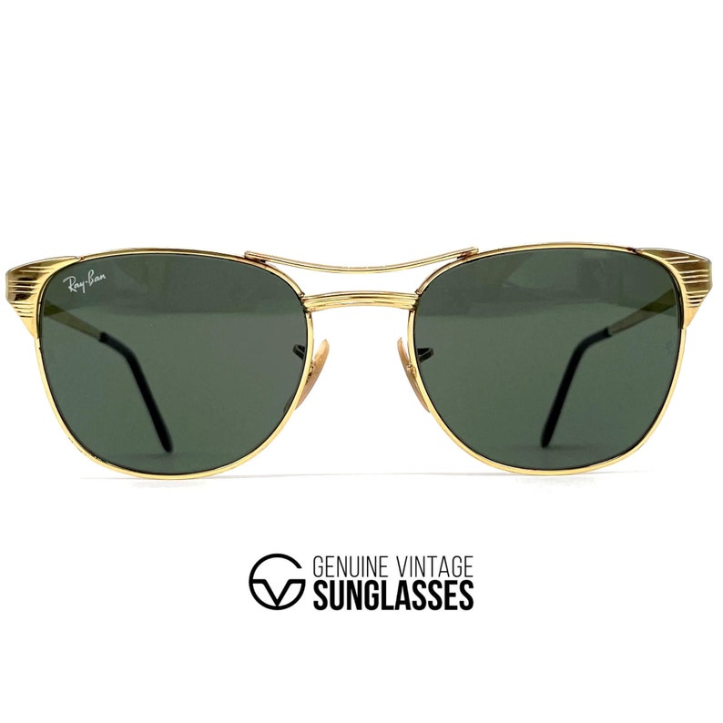 Op de afbeelding: Goudkleurige metalen zonnebril met een dubbele brug en donkergroene glazen. Het witte Ray-Ban-logo staat op het linkerglas. De randen van het montuur hebben een geribbelde textuur en de pootjes zijn dun met zwarte uiteinden. De tekst GENUINE VINTAGE SUNGLASSES staat eronder.