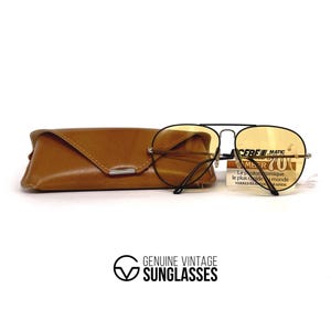 May include: Classic aviator sunglasses with amber lenses and a black frame, alongside a brown leather case. The glasses have a tag with text. The logo "GENUINE VINTAGE SUNGLASSES" is at the bottom.