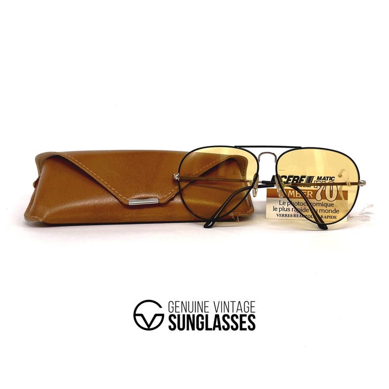 May include: Classic aviator sunglasses with amber lenses and a black frame, alongside a brown leather case. The glasses have a tag with text. The logo "GENUINE VINTAGE SUNGLASSES" is at the bottom.