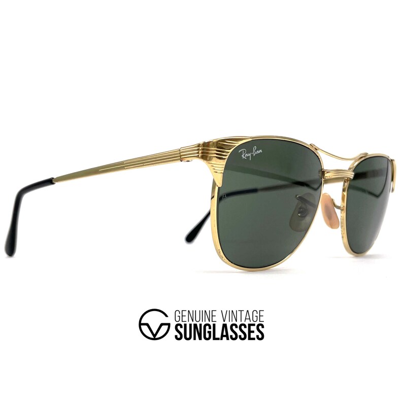 Op de afbeelding: Goudkleurige metalen zonnebril met donkergroene glazen en een textuur op het montuur. Het rechterglas is voorzien van het witte Ray-Ban logo. De pootjes zijn dun met geribbelde details en zwarte kunststof uiteinden. De neuspads zijn lichtbruin.