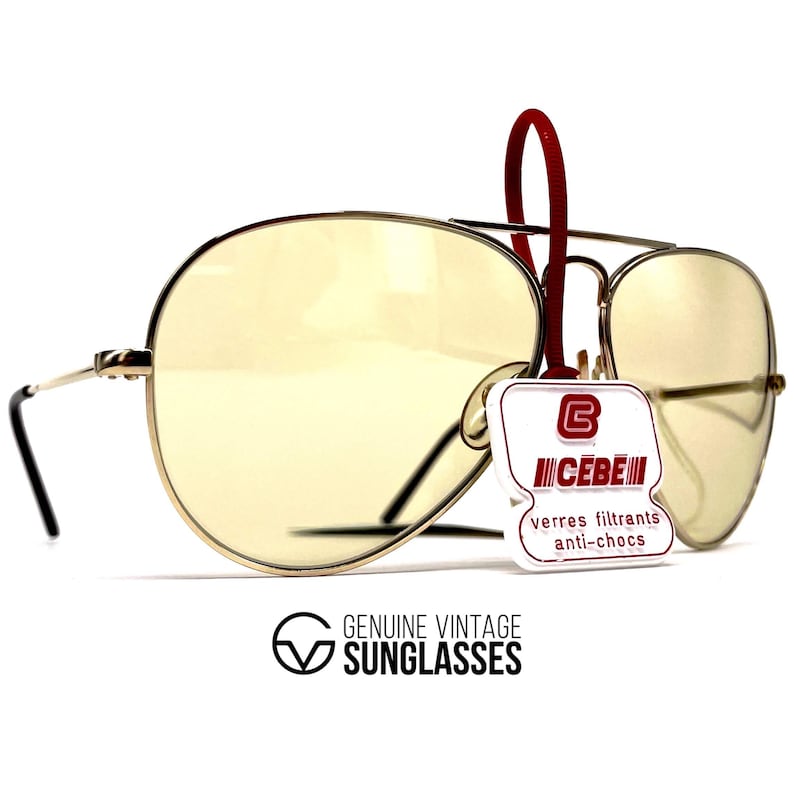 May include: Vintage aviator sunglasses with yellow-tinted lenses and a silver metal frame. The sunglasses have a red tag attached with the brand name "CEBE" and the text "verres filtrants anti-chocs". The text "GENUINE VINTAGE SUNGLASSES" is also visible.