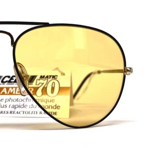 May include: Close-up of vintage sunglasses with yellow lenses and a black frame. The glasses have a classic aviator shape with a gold-coloured bridge and temples. A tag with text is visible behind the lens.