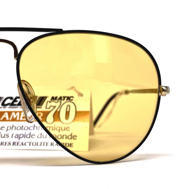 May include: Close-up of vintage sunglasses with yellow lenses and a black frame. The glasses have a classic aviator shape with a gold-coloured bridge and temples. A tag with text is visible behind the lens.