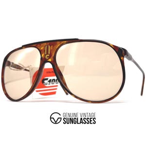 May include: Vintage Carrera C100 sunglasses with a tortoiseshell frame and light brown lenses. The sunglasses have a classic aviator shape with a double bridge and a tag attached. The text "GENUINE VINTAGE SUNGLASSES" is at the bottom.