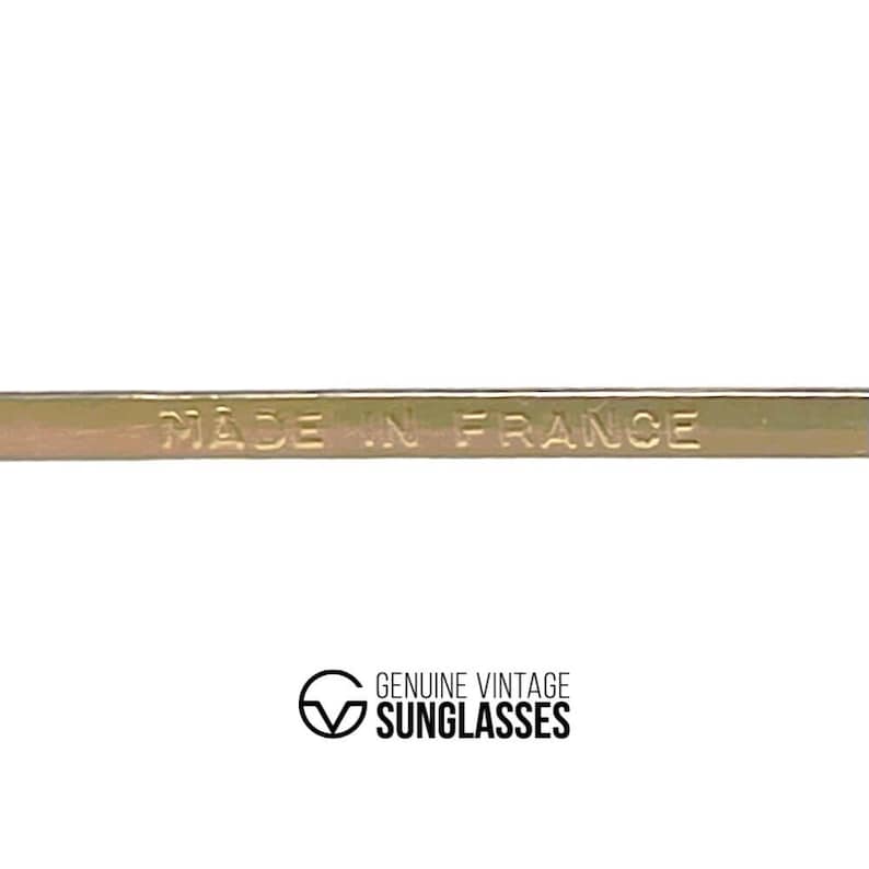 Puede incluir: Primer plano de una montura met&aacute;lica dorada con el texto en relieve "MADE IN FRANCE". Debajo, el logotipo dice "GENUINE VINTAGE SUNGLASSES" en una fuente serif. La montura es probablemente para gafas o gafas de sol.
