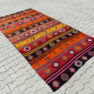 Pode incluir: Um tapete kilim turco vintage colorido com um padr&atilde;o geom&eacute;trico em tons de vermelho, laranja, amarelo, preto e rosa. O tapete tem um design listrado com motivos repetidos.