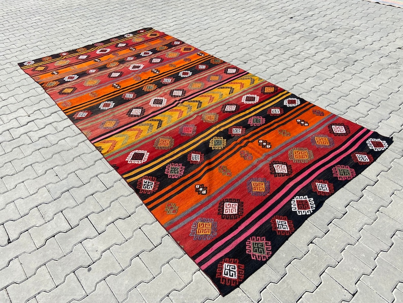 Pode incluir: Um tapete kilim turco vintage colorido com um padr&atilde;o geom&eacute;trico em tons de vermelho, laranja, amarelo, preto e rosa. O tapete tem um design listrado com motivos repetidos.