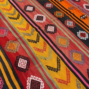 Pode incluir: Um tapete colorido com um padr&atilde;o geom&eacute;trico em tons de vermelho, laranja, amarelo, preto e branco. O tapete tem um design tradicional com quadrados e linhas repetidos.