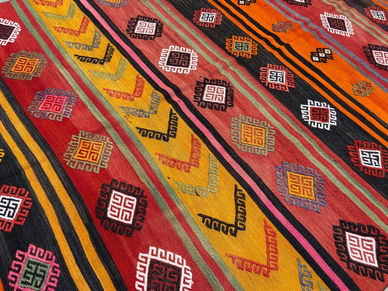 Pode incluir: Um tapete colorido com um padr&atilde;o geom&eacute;trico em tons de vermelho, laranja, amarelo, preto e branco. O tapete tem um design tradicional com quadrados e linhas repetidos.