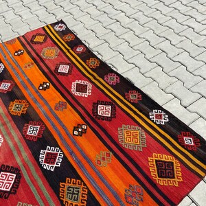 Pode incluir: Um tapete colorido tecido &agrave; m&atilde;o com um padr&atilde;o geom&eacute;trico em tons de vermelho, laranja, preto e branco. O tapete tem um design tradicional com motivos repetidos.