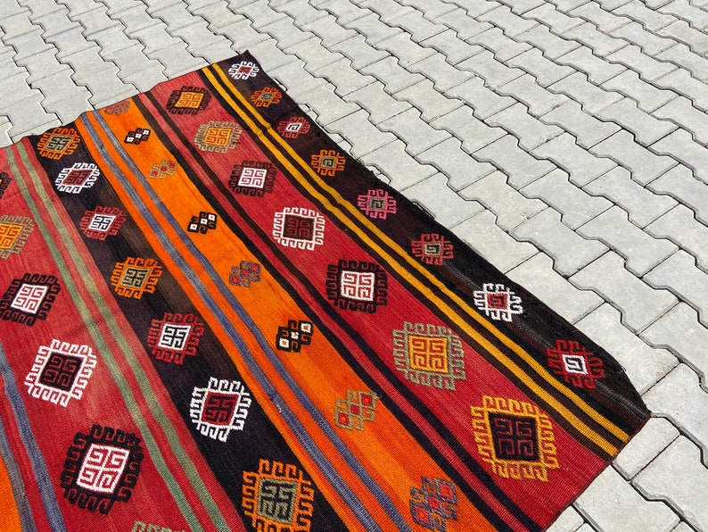 Pode incluir: Um tapete colorido tecido &agrave; m&atilde;o com um padr&atilde;o geom&eacute;trico em tons de vermelho, laranja, preto e branco. O tapete tem um design tradicional com motivos repetidos.