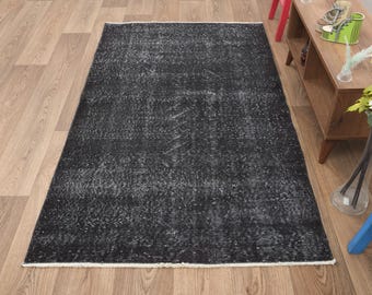 Puede incluir: Una alfombra rectangular negra desgastada con un borde blanco. La alfombra tiene una apariencia descolorida y vintage, con acentos grises claros y blancos en todo el campo negro. La alfombra está colocada sobre un suelo de madera clara.
