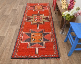 May include: A red, orange, and black patterned runner rug with a geometric design. The rug has a fringe border and is laid out on a wood floor.