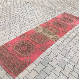 Puede incluir: Una alfombra larga y estrecha con fondo rosa y un patr&oacute;n geom&eacute;trico en tonos marrones y beige. La alfombra tiene flecos en ambos extremos.