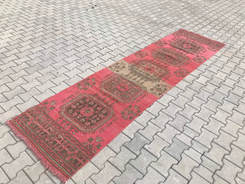 Puede incluir: Una alfombra larga y estrecha con fondo rosa y un patr&oacute;n geom&eacute;trico en tonos marrones y beige. La alfombra tiene flecos en ambos extremos.