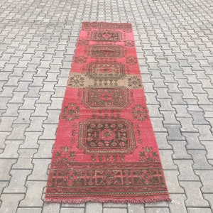 Puede incluir: Una alfombra larga y estrecha de color rosa y marr&oacute;n con un patr&oacute;n geom&eacute;trico. La alfombra tiene flecos en ambos extremos.