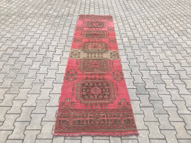 Puede incluir: Una alfombra larga y estrecha de color rosa y marr&oacute;n con un patr&oacute;n geom&eacute;trico. La alfombra tiene flecos en ambos extremos.