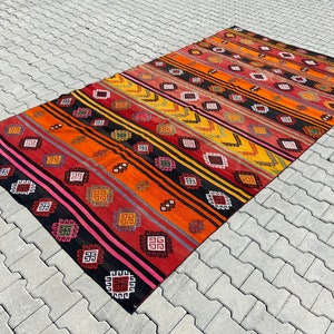 Pode incluir: Um tapete kilim turco vintage colorido com um padr&atilde;o geom&eacute;trico em tons de vermelho, laranja, amarelo, preto e rosa. O tapete tem um design listrado com motivos repetidos.