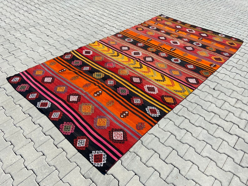 Pode incluir: Um tapete kilim turco vintage colorido com um padr&atilde;o geom&eacute;trico em tons de vermelho, laranja, amarelo, preto e rosa. O tapete tem um design listrado com motivos repetidos.