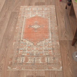 Puede incluir: Una alfombra de pasillo vintage con un centro naranja descolorido y un borde blanco y beige. La alfombra tiene un diseño persa tradicional con detalles intrincados.