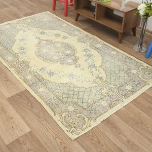 Puede incluir: Alfombra rectangular vintage con un campo central crema y un borde detallado. La alfombra presenta un patrón floral en tonos azules y grises, con un aspecto envejecido. La alfombra está colocada sobre un suelo de madera.