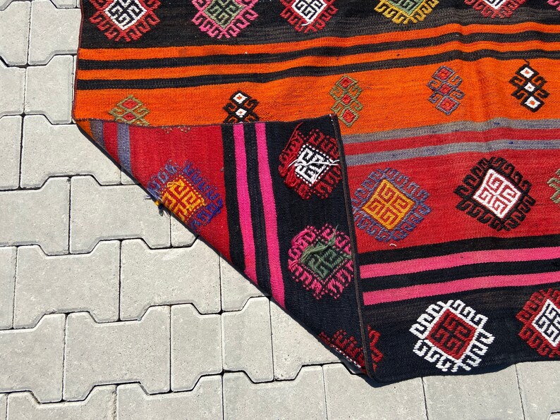 Pode incluir: Um tapete tecido colorido com padr&otilde;es geom&eacute;tricos em tons de vermelho, laranja, preto, rosa e azul. O tapete tem uma borda franjada e &eacute; feito de l&atilde;.